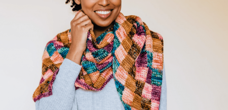 A Black woman smiles as she models a shawl made up of brightly colored squares