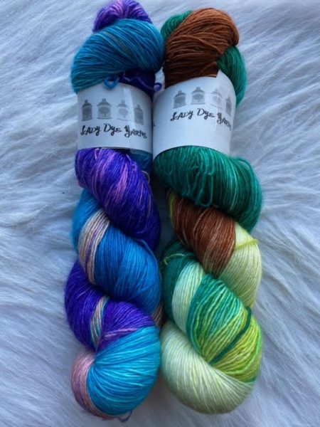 Two skeins of yarn on a white fur background. The skein on the left is made of purples and blues. The skein on the right is greens and browns.