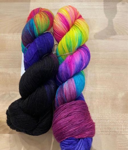Two skeins of yarn side by side. One is black with rainbow colors shot through. The other is a rainbow color with more purple in the base.