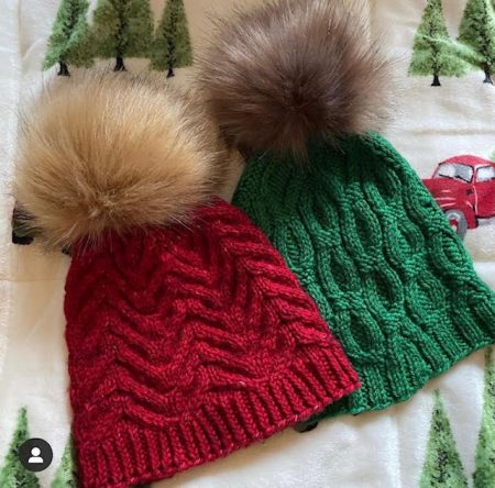 Two hats set against a Christmas blanket. One is red, one is green, and both have fake fur pom poms.