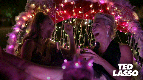 Keeley and Rebecca Bond while laughing over drinking entireb ottles of champagne under a lighted arch and feathers