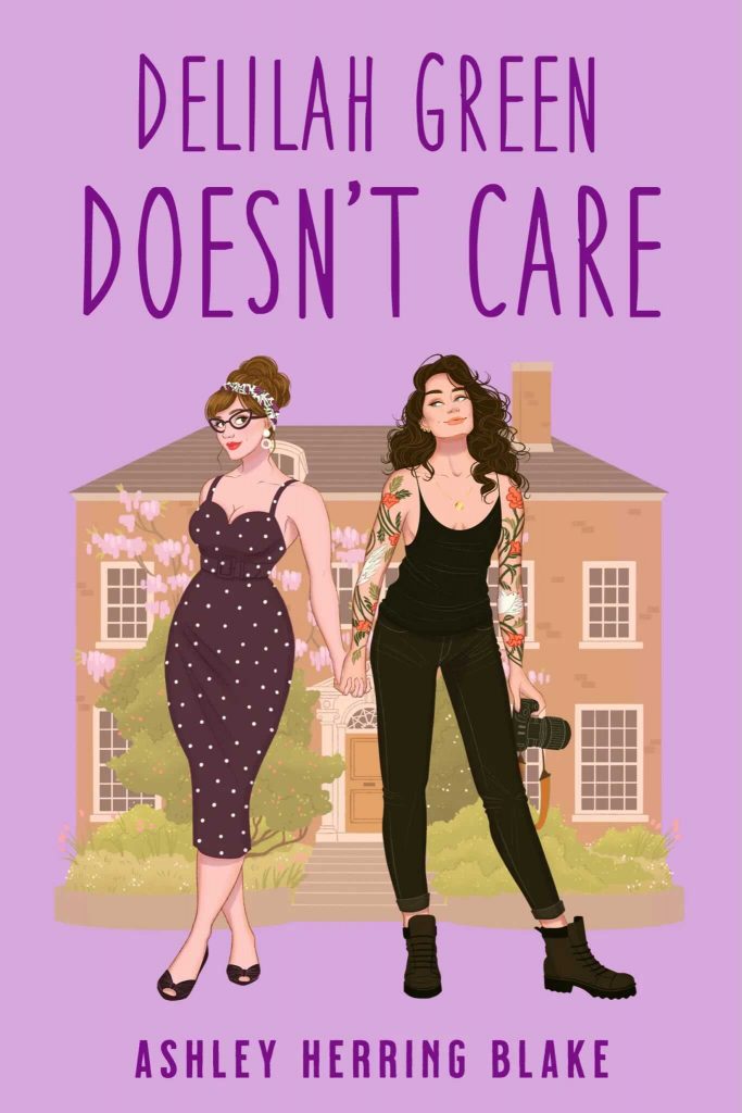 Delilah Green Doesn't Care by Ashley Herring Blake. Two women holding hands in front of a house. One has pinup vibes with bun tied with a scarf and body hugging dress. The other woman has wild wavy hair, is dressed in a black tank and jeans. Her arms are covered in tattoos.