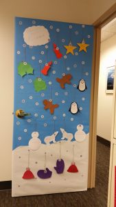 Maggie's awardwinning door with a blue background and a white hill of snow at the bottomw with cutouts of penguins, snow hats and gingerbread men hanging from the door each with a pun joke written on them!