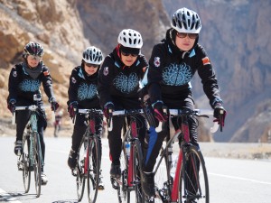 Women on cycling team biking up hill in Afghanistan. Their jackets have a light blue pattern on the chest, almost like a sun