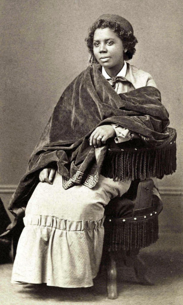 Edmonia Lewis, wearing a shawl and a dress with a ruffle at the hem, with a pillbox hat in a black and white photograph.