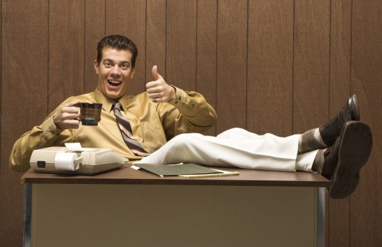 Truly repellant dude in a mustar dshirt against wood paneling giving a very smarmy thumbs up with his feet on a desk