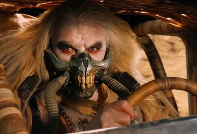 Immortan Joe, a light skinned man with long light crusty hair and a face mask that has tubes coming out of it. The facemask makes it look like he has massive teeth like a raptor or predator