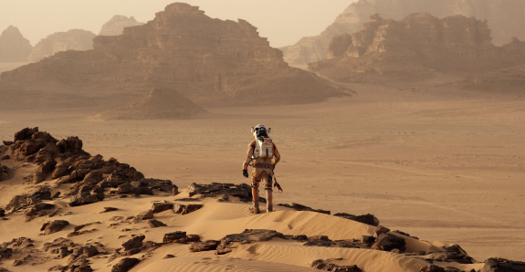 A shot of Matt Damon in a space suit walking on sand and rocks against a very red backdrop of Mars