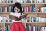 A little girl with glasses and a red skirt reading a big book in the library
