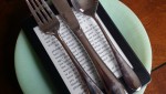 A fork knife and spoon resting on top of a Kindle, placed on top of a light green jade glass plate on a wood table - photo by Sarah Wendell
