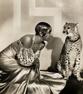 Josephine baker with gelled curls against her cheek in an off-the shoulder gown, smiling at a cheetah. The cheetah is unimpressed.