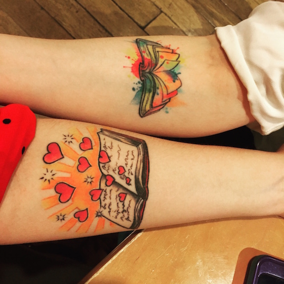 Close up of their forearms, each with a tattoo near the elbow joint. Elyse is a book with paint splattered on top and Amanda's is a book with hearts coming out of the pages and rising into the air above the book
