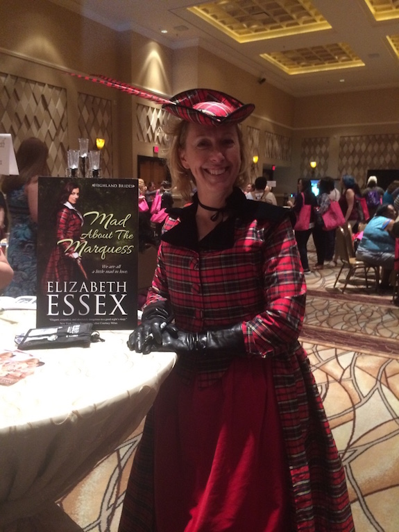 Half-body shot of Elizabeth Essex in a red plaid jacket with a dark collar. The jacket reaches to the floor like a dress, and she's wearing a red fascinator-style hat with a sharp red feather sticking out. 