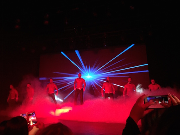 Shot of the performers in red smoke with a laser backdrop HELLO THE 80S each wearing tight pants and a bowtie and sporting a LOT of musculature.