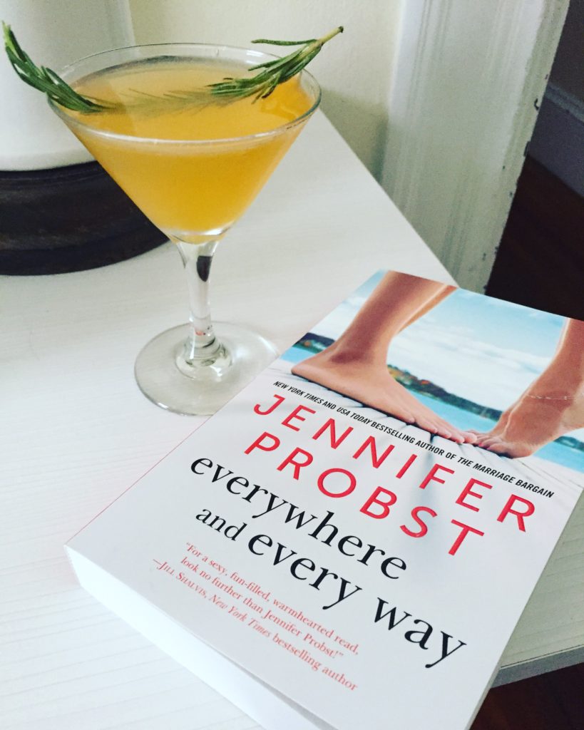 The Sweet Hot Chris (a bright yellow drink) in a cocktail glass with a sprig of rosemary and a copy of Everywhere and Every Way
