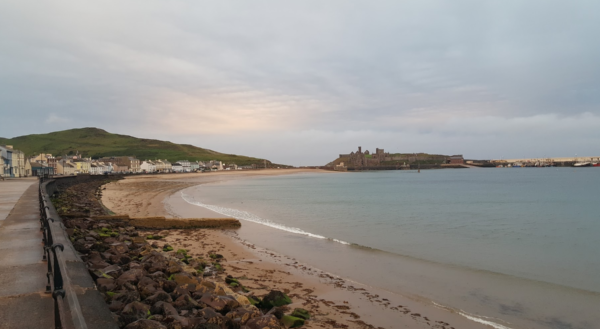 A long curving stretch of beach under a peach and blue sky with the medival castle in the distance