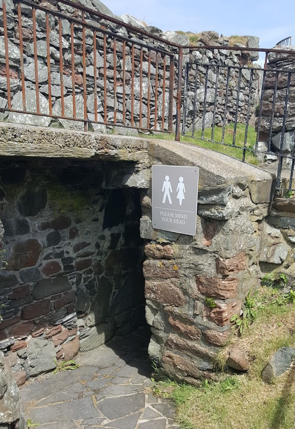 A placard reads PLEASE MIND YOUR HEAD Beneath a male and female figure designating a bathroom or WC. Behind the sign is a tunnel which leads to a turret where I kid you not the bathrooms have been installed