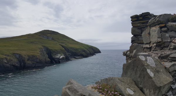 The grey rock of the castle on the right with the inlet of the sea in front of it and the green rise of Peel Hill beyond the water