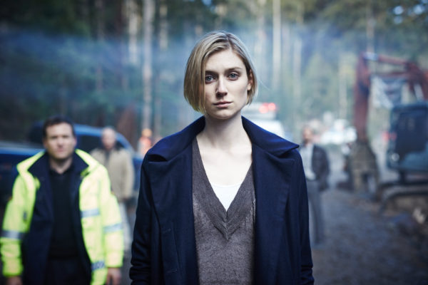 Anna who is pale wearing a grey shirt and blue coat with chin length straight blonde hair in a bob to her chin looking at the camera with a wan pallid expression