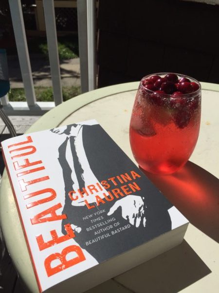 A copy of Beautiful by Christina Lauren next to a glass of fall sangria on a table in the sun.