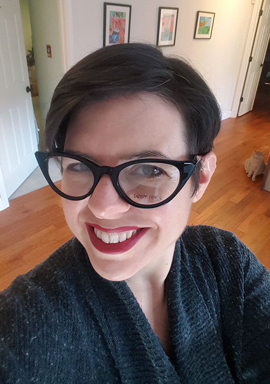 Me wearing black cats eye glasses with a goofy smile on my face and orville, my cat, in the hallway visible over my shoulder wondering what the hell I am doing. 