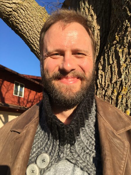 Rich is standing in front of a tree, wearing his cowl unbuttoned