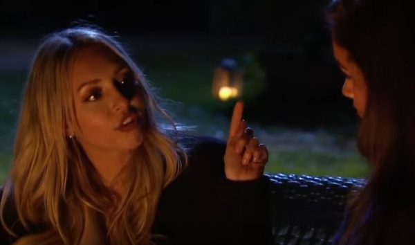 Corinne sits facing Taylor on a couch. She's holding up her finger, arguing. 