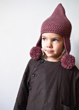 A youn girl wears a pointed hat with ear flaps and pom poms on the end of the flaps. 