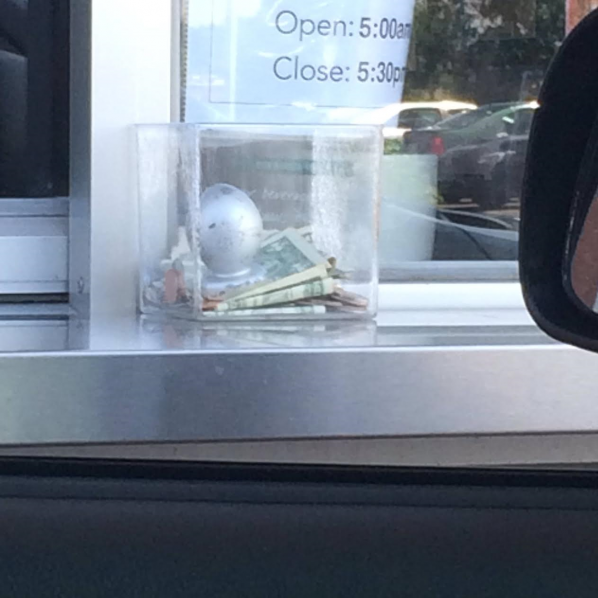 A silver object (possibly butt plug, possibly newel post cap) sits on top of a pile of cash in a clear box outside a Starbucks drive through.