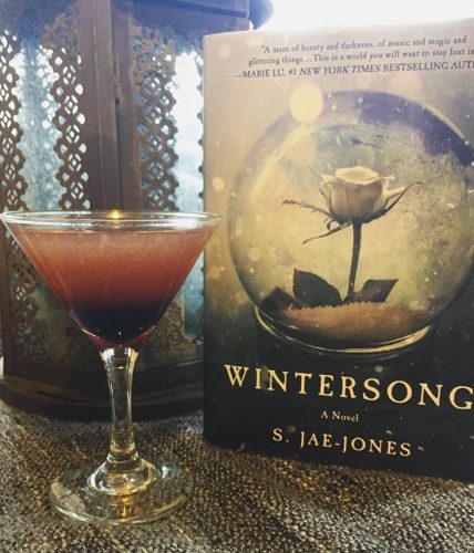 A pink and purple cocktail next to a copy of Wintersong and a rusty, old lamp.