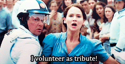 Katniss from the Hunger Games rushes forward through a crowd while shouting "I volunteer as tribue!"