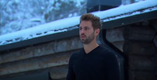 Nick is standing outside a snow covered cabin wearing a sweater and staring into the middle distance.