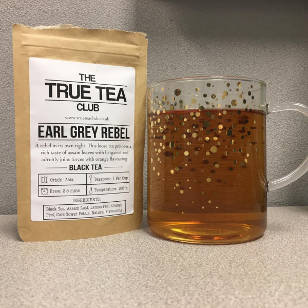 The pouch of Earl Grey Rebel sits next to my glass tea cup filled with delicious tea. The background is my ugly gray desk at work.