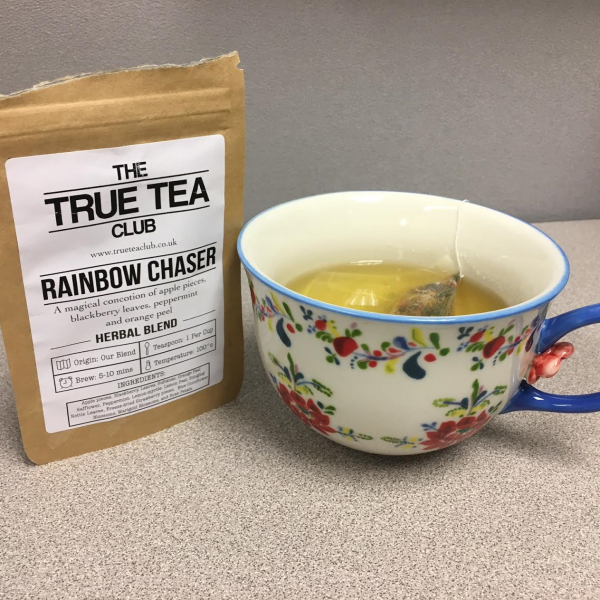 The package of Rainbow Chaser tea sits next to a delicate, floral teacup filled with tea. My desk is still gray and ugly.