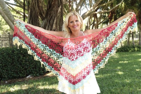 A smiling woman holds up a large, lacey triangluar shawl crocheted in pink, blue, red and yellow.