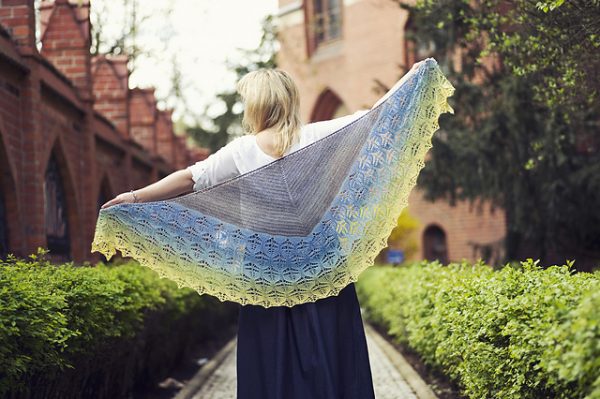 A woman models a crescent-shaped shawl that starts off grey then has leaf-patterned lace work in a blue to yellow gradient.