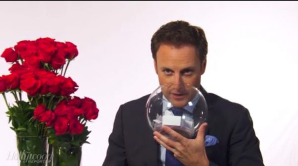 Chris Harrison is holding a glass bowl up to his face while sitting next to a bunch of roses.