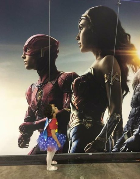 A very small girl in a wonder woman tutu looking up at a movie poster of gal godot as ww