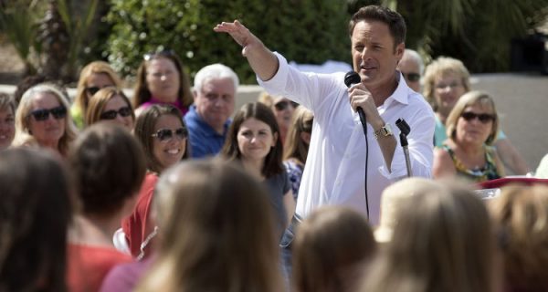 Chris Harrison stands in the middle of a crowd, talking into a mic.