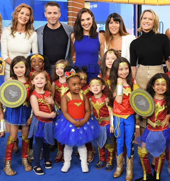 The cast and director surrounded by small girls of different heights and colors all dressed as wonder woman.