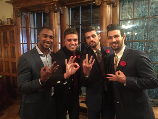 Four contestants stand next to each other in suits, making dumb hand gestures and faces.