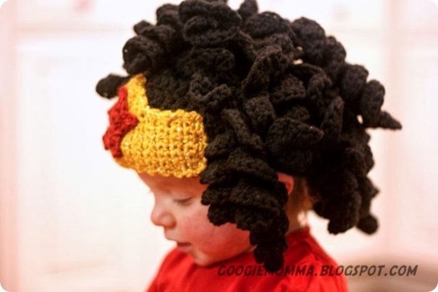 A super adorable baby wears a hat that is crocheted to look like Wonder Woman's headband with her long black curls flowing out from over it.