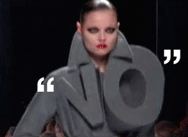 Model on runway wearing grey coat that spells out in three dimensional letters NO 