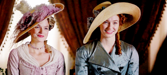 Caro and D, side by side, in fancy clothes and wearing shit-eating grins as they charm men out of their money.