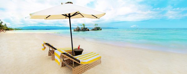 A white sand beach with a calm, turquoise ocean. Two lounge chairs face the ocean, an umbrella keeping them in the shade. Between them is a table with a tropical drink on it.