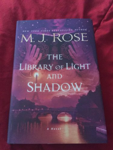The Library of Light and Shadow by MJ Rose. A woman holding a book and the scene of the Eiffel Towel is awash in varying shades of rose and purple.