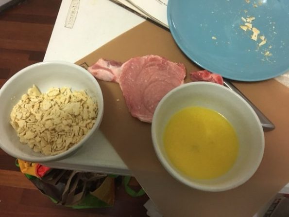 Crushed crackers in a bowl, a smashed pork chop, and melted butter with a beaten egg in another bowl