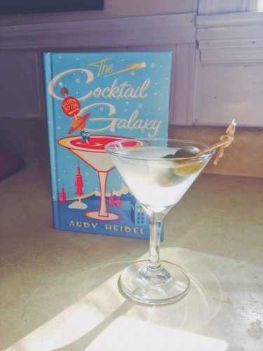 A martini glass with a clear drink, garnished with olives. The Cocktail galaxy is positioned behind the drink