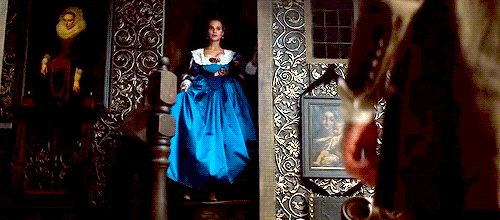 Alicia Vikander coming down into a room with patterned walls (leaves in curly-cue shape all over the wall) wearing a blue gown with a white lace collar.