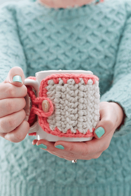 A woman with a seriously gorgeous manicure holds a mug wrapped in a pink and ivory mug cozy.